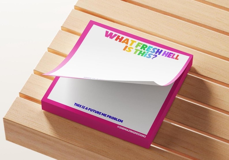 May include: A pink notepad with the text "WHAT FRESH HELL IS THIS?" in rainbow colours and "THIS IS A FUTURE ME PROBLEM" in blue. The notepad is on a wooden surface.