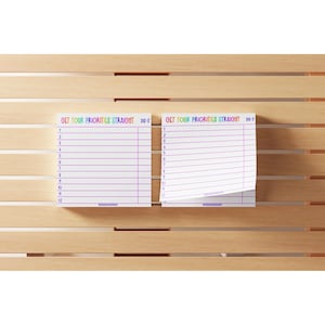 May include: Two white notepads with the text "GET YOUR PRIORITIES STRAIGHT DID IT" in rainbow colors. Each notepad has 12 numbered lines for listing priorities. The notepads are on a wooden surface.