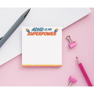 May include: White notepad with the text "ADHD IS MY SUPERPOWER" in blue, orange, and yellow. The notepad has a decorative orange and pink border. A gray pen, pink pencil, and pink binder clips are also visible.