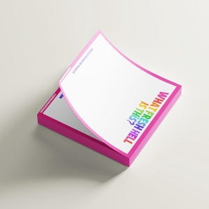 May include: A pad of pink sticky notes, featuring the text "WHAT FRESH HELL IS THIS?" in rainbow hues. The top sheet is lifted, showing the white paper beneath. The edges of the notes are a bright pink colour.