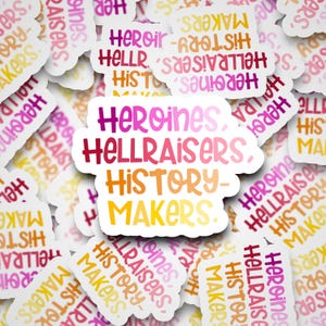 May include: A collection of white stickers with the words "Heroines, Hellraisers, History-Makers" in a gradient of pink, red, orange, and yellow. The stickers are scattered across a white surface.