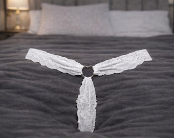May include: White lace lingerie with a heart-shaped metal ring. The delicate lace is arranged in a T-shape, with a scalloped edge. The lingerie is displayed on a dark grey bedspread.