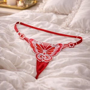 Op de afbeelding: Rode string met een vlinderontwerp. Het lingerie heeft een rode stof met een vlinderapplicatie in rood en wit. De verstelbare bandjes hebben bloemaccenten. De string wordt gepresenteerd op een wit bed.