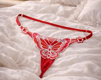 May include: Red thong lingerie with a butterfly design. The underwear features a red fabric with a butterfly applique in red and white. The adjustable straps have floral accents. The lingerie is displayed on a white bed.