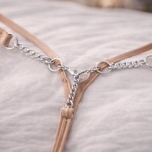 Puede incluir: Un tanga beige con detalles de cadena plateada. La tanga presenta finas correas y un diseño de cadena que conecta la parte delantera y trasera. El fondo es una tela blanca y suave.