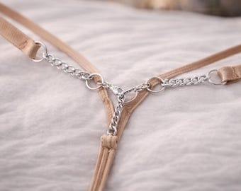 May include: A beige thong with silver chain detailing. The lingerie has thin straps and a chain design connecting the front and back. The background is a soft, white fabric.