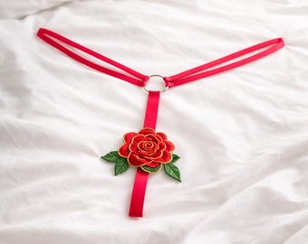 May include: Red lingerie with a floral accent. The lingerie features a red ribbon design with a silver ring and a red rose with green leaves. The lingerie is on a white bed.
