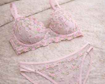 May include: A light pink bra and matching briefs set. The lingerie features delicate floral embroidery in shades of pink and green. The set is displayed on a bed with a textured, light-coloured comforter.