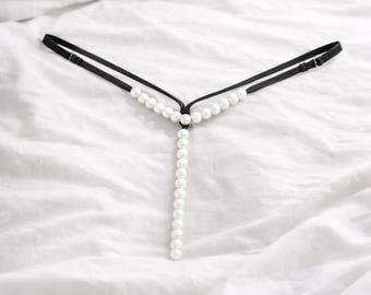 May include: Black lingerie with a pearl detail. The lingerie has a black elastic band and a vertical strand of white pearls. The garment is displayed on a white bedsheet.