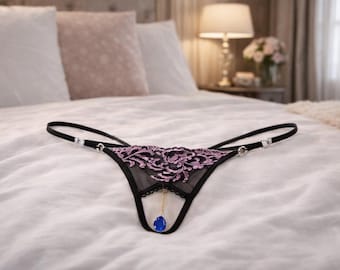 May include: Black lingerie with a floral lace design in shades of purple and black. The lingerie features a blue teardrop-shaped charm and rhinestone accents. The garment is displayed on a white bed.