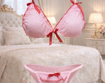 May include: A light pink lingerie set comprising a triangle bra and a thong. Both items feature red ribbon detailing. The bra has red trim and bows. The lingerie is made of a soft, silky material.