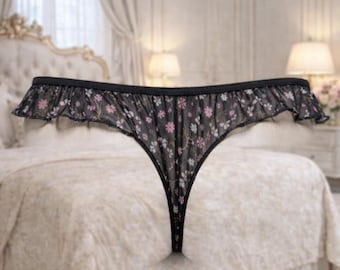 May include: Black lingerie with a floral pattern in pink and white. The lingerie has a ruffled edge and a thong back. The garment is displayed against a neutral background.