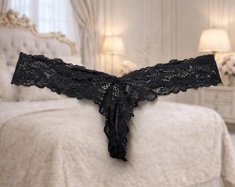 May include: Black lace thong. The underwear is made of delicate floral lace and has a low-rise cut. The thong is displayed on a bed with a cream-coloured comforter and a ruffled bed skirt. The background includes a chandelier and lamps.