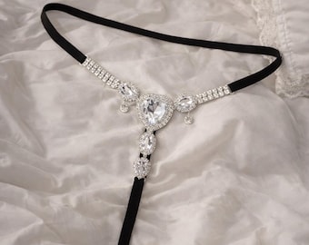 May include: A black elastic headpiece featuring a central heart-shaped rhinestone design and dangling rhinestones. The headpiece has a black vertical strap with additional rhinestones. The background is a white, textured fabric.