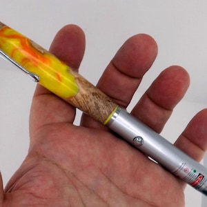 Op de afbeelding: Een laserpointer met een zilveren behuizing en een kleurrijke handgreep. De handgreep heeft een geel en oranje wervelpatroon en een houtnerfpatroon. Een zilveren clip is bevestigd. Ongeveer 15 cm lang.