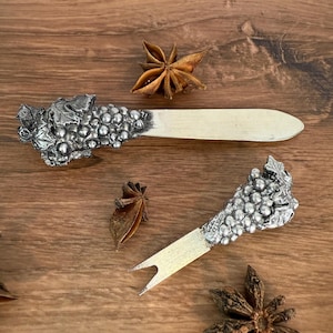 May include: Two silver-toned cheese spreaders with grape cluster designs on the handles. One is a knife, the other a fork. The spreaders are on a wooden surface, with star anise scattered around.
