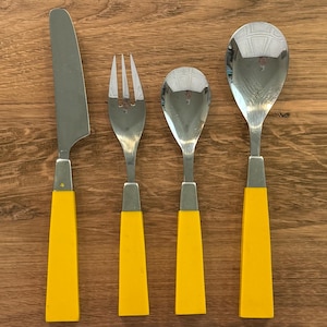 May include: A set of four stainless steel utensils with bright yellow handles. The set includes a knife, fork, and two spoons. The utensils are arranged on a wooden surface.