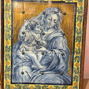 Pode incluir: Obra de arte em azulejo emoldurada retratando uma cena religiosa. A obra apresenta uma mãe e uma criança em tons de azul e branco, contra um fundo dourado. A moldura é de madeira escura e os azulejos são dispostos em grade.