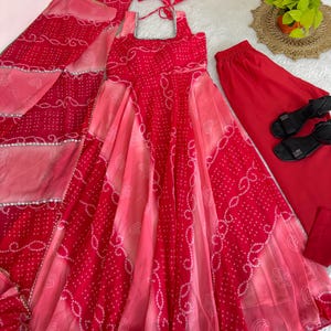 May include: A vibrant red and pink Indian salwar kameez set. The set includes a long dress, pants, a scarf, and a matching headband. The dress and scarf feature a white dotted pattern. Black sandals are also included.