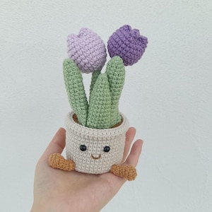 Peut inclure: Un pot de plante crocheté avec un visage souriant, tenant deux tulipes violettes et lavande. Le pot est beige avec des pieds marron. Les tiges et les feuilles vertes ajoutent une touche de nature à cet article fait à la main.