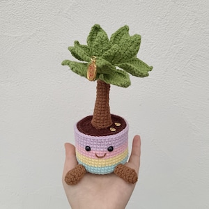 Può includere: Una palma all'uncinetto con un volto sorridente sul vaso. L'albero ha foglie verdi, un tronco marrone e un ciondolo dorato. Il vaso è a righe con colori pastello: viola, giallo e blu. Un articolo fatto a mano.