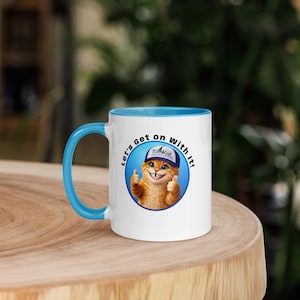 May include: White ceramic mug with a light blue handle and interior. The mug features a cartoon cat wearing a hat and giving a thumbs-up, with the text "Let's get on with it!"