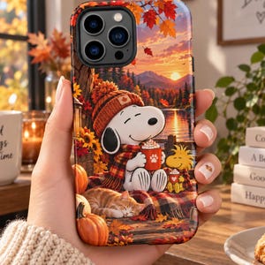 May include: A smartphone case featuring a colorful autumn scene with Snoopy wearing a knit hat and scarf, holding a mug next to Woodstock and a sleeping orange tabby cat. The background shows a sunset over a lake with mountains and falling leaves.