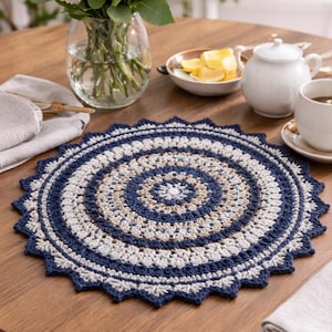 May include: A round, crocheted doily in navy, cream, and beige tones, with a decorative scalloped border. The doily sits on a wooden table, with a vase of white flowers, a teapot, and a cup of tea nearby.