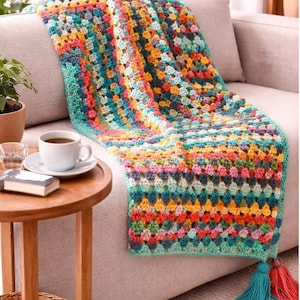 Puede incluir: Una colorida manta de ganchillo hecha a mano, sobre un sofá beige. La manta presenta una vibrante mezcla de cuadrados turquesa, naranja, amarillo y rosa. Dos borlas en turquesa y rojo cuelgan del borde. Una taza de café está sobre una mesa auxiliar.