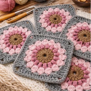 May include: Five crocheted square coasters with a floral design. Each coaster has a brown centre, surrounded by pink and light pink petals, and a grey border. Yarn and crochet hooks are visible in the background.