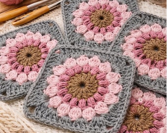 May include: Close-up of five crocheted square coasters. Each coaster features a floral design with a brown center, surrounded by pink and light pink petals, and a gray border. Yarn and crochet hooks are visible in the background.