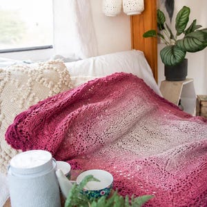 May include: A handmade, crocheted blanket in shades of pink and white, draped over a bed. The blanket features a scalloped edge and an open, lacy design. Decorative items like a white jar, cups, and a plant are also visible.