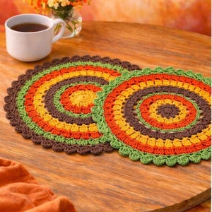 May include: Two handmade, circular crochet placemats in warm autumn colors: brown, orange, yellow, and green. The placemats are placed on a wooden table, with a cup of tea and a vase of flowers in the background. The crochet design features a scalloped edge.