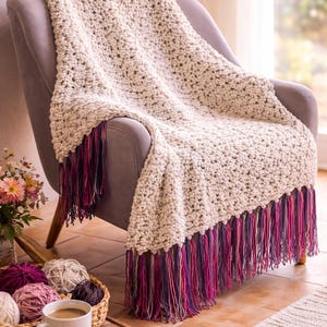 May include: A cream-colored, chunky knit throw blanket with a fringe of purple, pink, and burgundy yarn is draped over a gray armchair. The blanket is handmade and adds a cozy touch to the room.