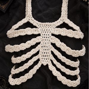 May include: A cream-coloured crochet top featuring a rib cage design. The garment is constructed with a central vertical panel and horizontal bands extending outward to mimic the appearance of human ribs, finished with two shoulder straps.