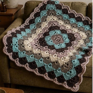 Op de afbeelding: Een handgemaakte gehaakte deken in een cirkelvormig patroon, met een mix van kleuren waaronder teal, bruin, beige en lavendel. De deken is over een groene bank gedrapeerd, wat het ingewikkelde ontwerp en de textuur laat zien. De afmetingen van de deken zijn niet zichtbaar.