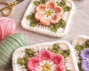 May include: Handmade crochet coasters with floral designs. Each square coaster features a white background with green leaves and a central flower in shades of pink, peach, and yellow. Yarn and scissors are also visible.