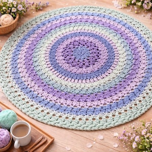 Puede incluir: Una alfombra redonda hecha a mano con ganchillo, en colores pastel. La alfombra presenta círculos concéntricos de lavanda, azul claro y verde menta. El centro tiene un patrón floral detallado. La alfombra tiene aproximadamente 122 cm de diámetro.