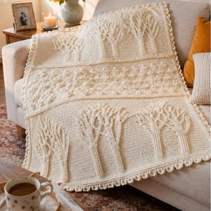 May include: Cream-colored crocheted blanket with a tree design. The blanket features rows of textured trees and a scalloped edge. The blanket is draped over a beige sofa, creating a cozy and inviting aesthetic.