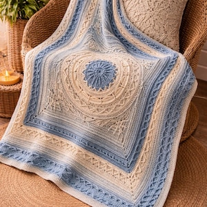 以下が含まれることがあります： クリーム色と青色の色合いの、かぎ針編みのブランケット。籐の椅子に掛けられています。ブランケットは中央に円形のデザインと複雑なボーダーパターンが特徴です。ブランケットのサイズは約152cm四方です。