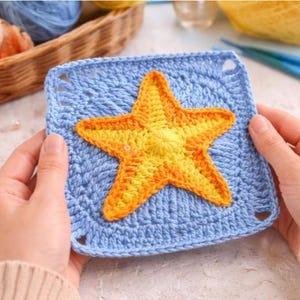 May include: A handmade crochet square in light blue with a vibrant yellow and orange starfish applique. The square has a textured stitch pattern and small corner holes. The starfish is centered on the square.