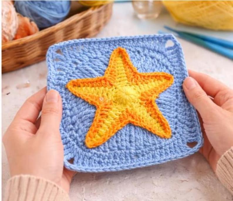 May include: A handmade crochet square in light blue with a vibrant yellow and orange starfish applique. The square has a textured stitch pattern and small corner holes. The starfish is centered on the square.