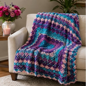 Op de afbeelding: Een kleurrijke, gehaakte deken in tinten paars, turquoise en roze ligt over een beige bank. De deken heeft een gedetailleerd, geometrisch patroon en een geschulpte rand. Een vaas met roze bloemen en een brandende kaars zijn zichtbaar.
