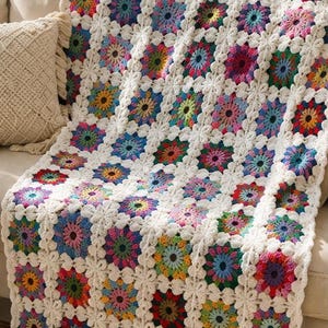 Può includere: Una coperta all'uncinetto fatta a mano e colorata, drappeggiata su un divano. La coperta presenta un motivo ripetuto di quadrati vibranti a forma di fiore nelle tonalità di blu, verde, giallo, rosa e rosso, bordati da uncinetto bianco. La coperta aggiunge un tocco accogliente e decorativo.