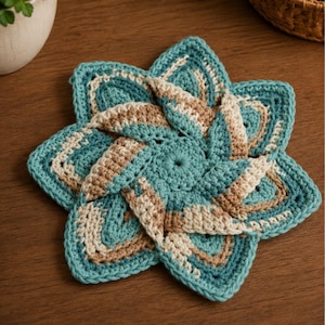 May include: A handmade, crocheted coaster in a starburst design. The coaster features a teal, tan, and cream color scheme. The coaster is made of yarn and is placed on a wooden surface. The coaster is a decorative item.