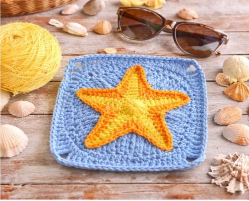 May include: A crocheted blue square with a yellow and orange starfish in the center. The image also includes a ball of yellow yarn, seashells, and sunglasses, all arranged on a wooden surface.
