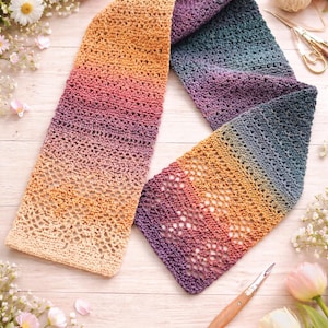 May include: A handmade, open-weave scarf in a gradient of colors, from light beige to deep purple. The scarf is crocheted with a delicate pattern and is surrounded by flowers and craft supplies.