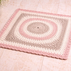 May include: A square, handmade crochet rug with a circular pattern in shades of pink, white, and gray. The rug has a scalloped pink border and is displayed on a light wood surface. The rug is approximately 24 inches square.