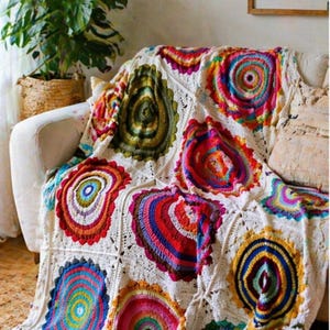 Puede incluir: Una colorida manta de ganchillo hecha a mano sobre un sofá blanco. La manta presenta grandes diseños circulares en tonos vibrantes de rojo, naranja, amarillo, azul y verde, creando una estética bohemia. Un acogedor artículo de decoración para el hogar.