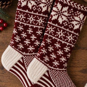 Puede incluir: Dos calcetines navideños de punto con un diseño en color burdeos intenso y crema. El patrón incluye copos de nieve detallados en el puño y la pierna, con un talón a rayas y un diseño geométrico texturizado en la zona del pie.